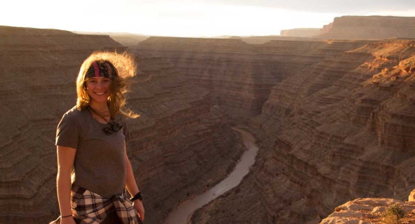 A person smiles as they stand high above a river framed by tall, red canyon walls. 